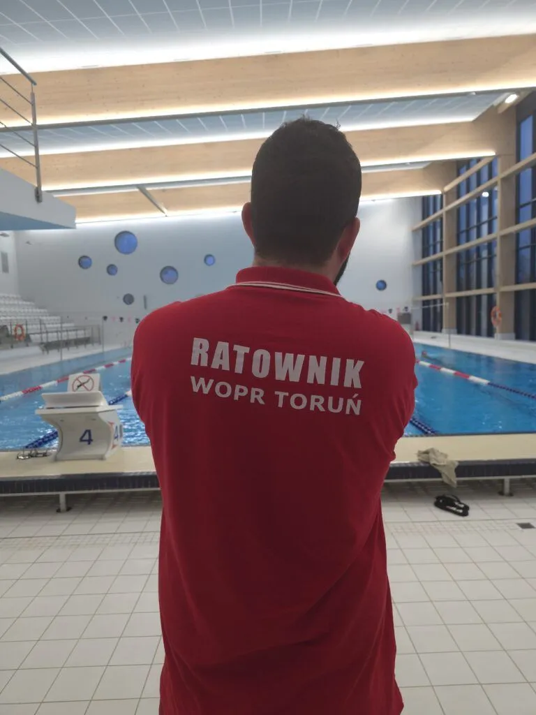 Ratownik w czerwonej koszulce z napisem Ratownik WOPR Toruń, widziany od tyłu, pilnuje krytej pływalni